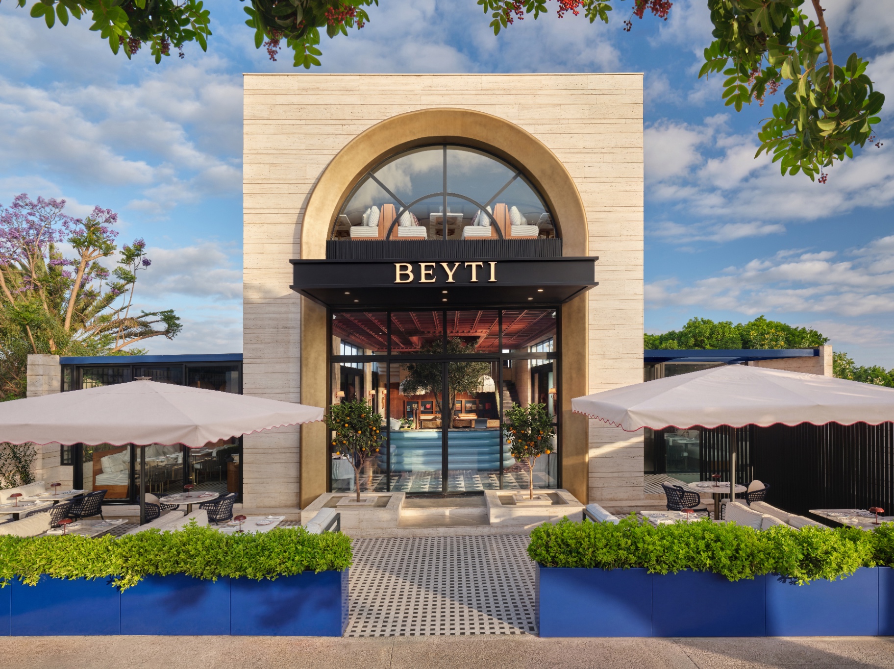 Beyti villa facade in Rabat with travertine arch, BEYTI signage, fringed parasols, and jacaranda blooms